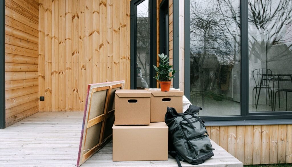 Wooden porch with cardboard boxes and potted plant next to glass windows.
